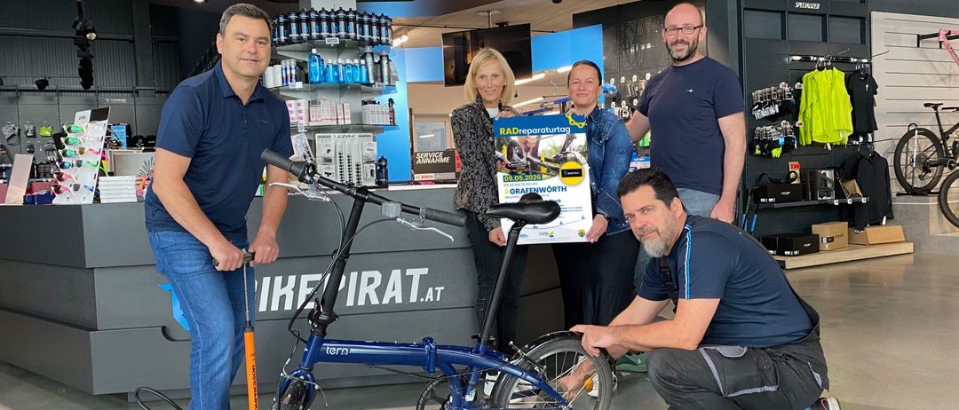 Eine Gruppe von Menschen posiert mit einem blauen Fahrrad in einem Geschäft. Ein Mann hält eine Pumpe. Hinter ihnen steht ein Regal mit verschiedenen Artikeln und einem Banner.