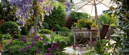 Ein Garten mit lila Blumen, einem Holztisch und Stühlen unter einem weißen Sonnenschirm.
