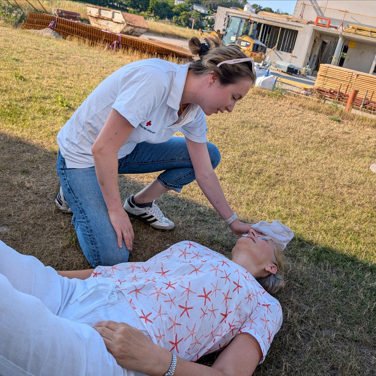 Eine Frau in einem weißen Shirt kniet auf einem Feld und kümmert sich um eine andere Person, die mit einem Tuch im Gesicht auf dem Boden liegt. Hinter ihnen befindet sich eine Baustelle mit einem Gebäude im Bau.