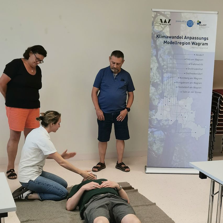 Drei Personen befinden sich in einem Raum mit einem Banner, das 'Klimawandel Anpassungs Modellregion Wagra' anzeigt. Ein Mann liegt auf einem Teppich, während eine Frau kniet und eine andere steht, beide mit Brille.