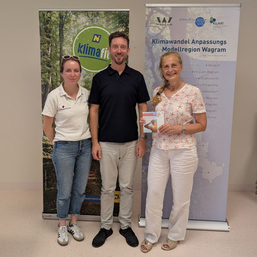 Drei Personen stehen vor einem Hintergrund mit dem Klimafit-Logo. Eine Frau links trägt ein weißes Poloshirt und blaue Jeans, während der Mann in der Mitte ein schwarzes Poloshirt und Khakihosen trägt. Eine andere Frau rechts hält ein Buch und lächelt.