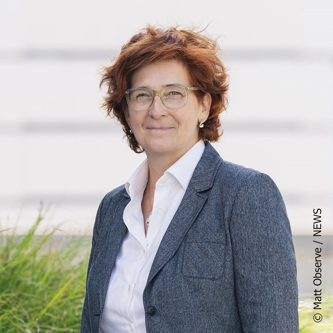 A woman with short red hair and glasses is wearing a gray jacket and a white shirt. She is standing outside.