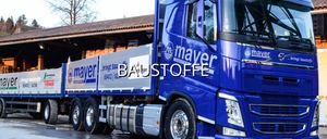 Ein blauer Lkw mit der Aufschrift 'BAUSTOFFE' und dem Logo von Mayer Gunter steht vor einem Gebäude. Der Lkw hat einen großen Metallbehälter befestigt.