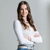 Eine Frau in einem weißen Langarmshirt und blauen Jeans steht mit verschränkten Armen und lächelt in die Kamera. Sie trägt eine Halskette und einen beigen Gürtel.
