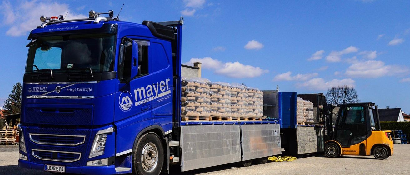 Ein blauer Lkw mit dem Wort Mayer an der Seite ist geparkt. Der Lkw transportiert einen Stapel Säcke mit Zement.