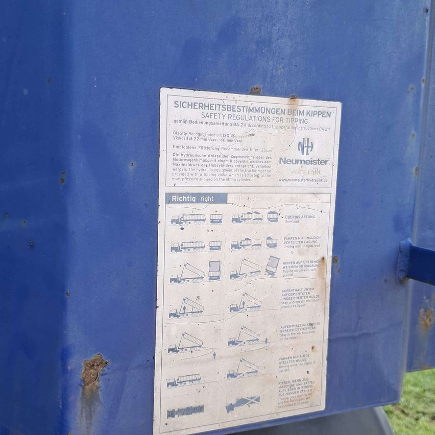 Ein blaues Schild bietet Sicherheitsbestimmungen für das Kippen auf Englisch und Deutsch. Es enthält Abbildungen von Maschinen und Anweisungen für den sicheren Betrieb.