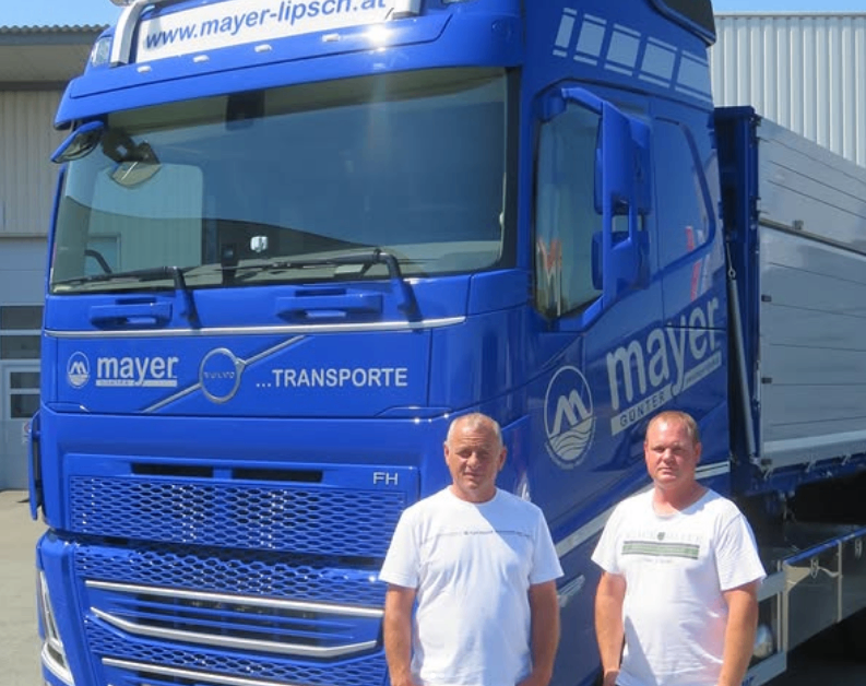 Zwei Männer stehen vor einem blauen Mayer-Lkw, beide tragen weiße T-Shirts. Der Lkw hat 'Transporte' und das Mayer-Logo auf seiner Vorderseite.
