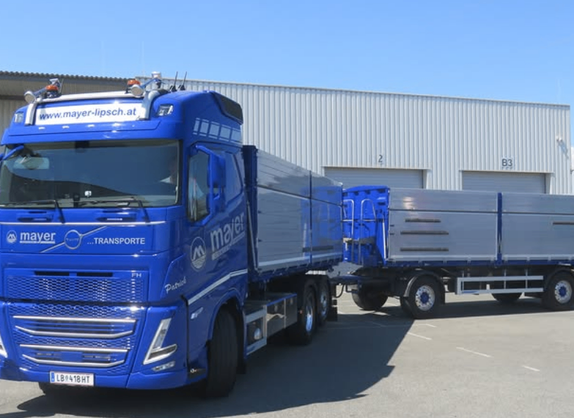 Ein blauer Lkw mit der Aufschrift 'Mayer Transport' steht vor einem Lager mit zwei Toren. Der Lkw hat einen großen Anhänger.