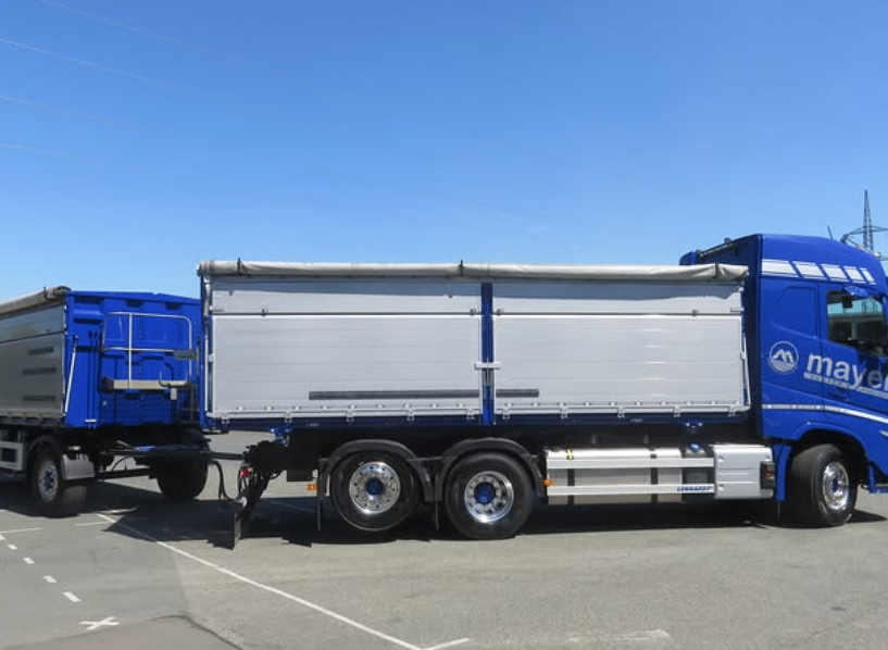 Ein großer blauer Sattelschlepper mit einem grauen Anhänger ist auf einer grauen Straße geparkt. Der LKW hat eine blaue Kabine mit einer Leiter an der Seite.