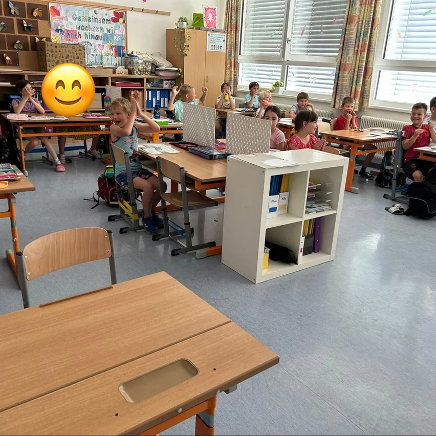 Ein Klassenzimmer mit Kindern an Tischen, ein lächelndes Kind hebt die Hände und ein fröhliches Emoticon oben. Die Tische sind in Reihen angeordnet, mit Regalen und einem weißen Bücherregal dazwischen.
