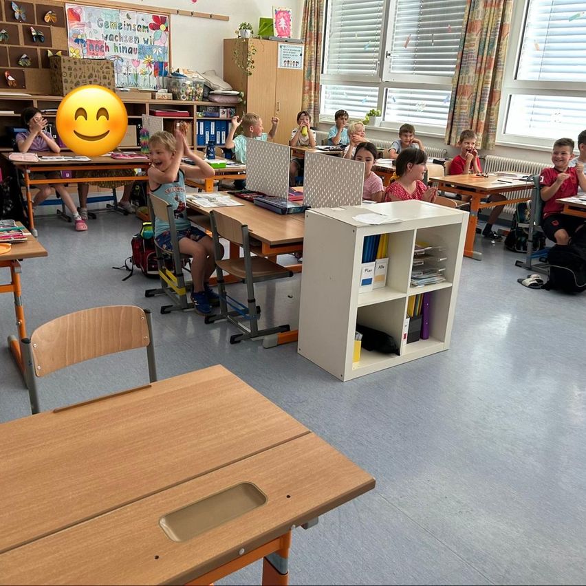 Ein Klassenzimmer mit Kindern an Tischen, ein lächelndes Kind hebt die Hände und ein fröhliches Emoticon oben. Die Tische sind in Reihen angeordnet, mit Regalen und einem weißen Bücherregal dazwischen.