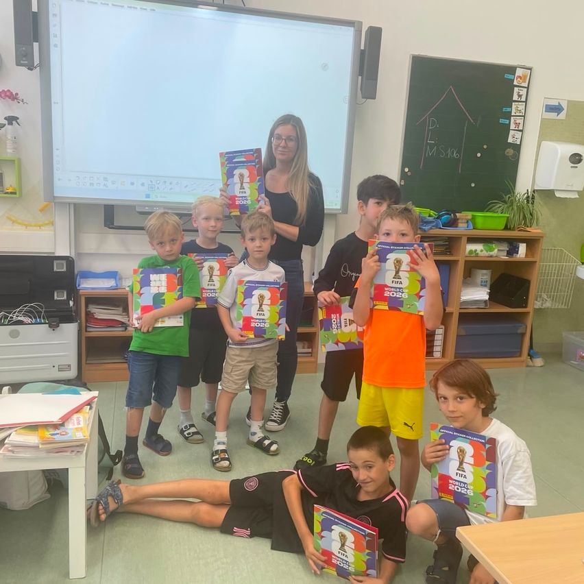Eine Gruppe von Kindern in einem Klassenzimmer posiert für ein Foto mit ihren Büchern. Eine Lehrerin steht hinter ihnen. Ein Whiteboard, Regale und eine Tafel sind im Hintergrund.