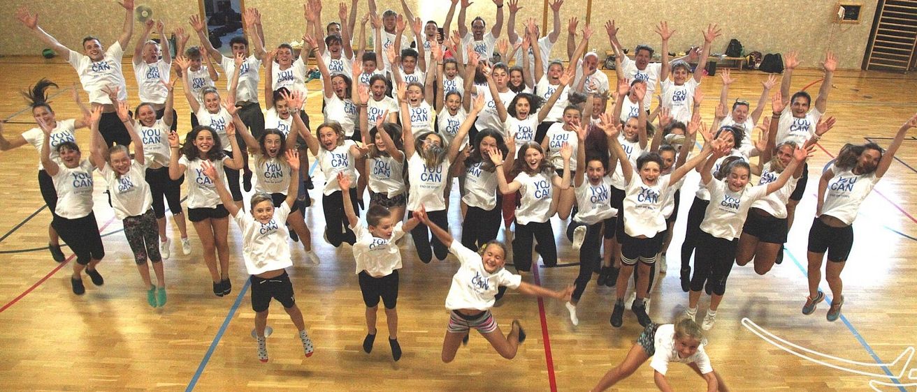 Eine Gruppe junger Menschen und Erwachsener, die weiße T-Shirts mit 'DU KANNST' tragen, posiert für ein Foto auf einem Basketballplatz. Sie springen und einige liegen auf dem Boden.
