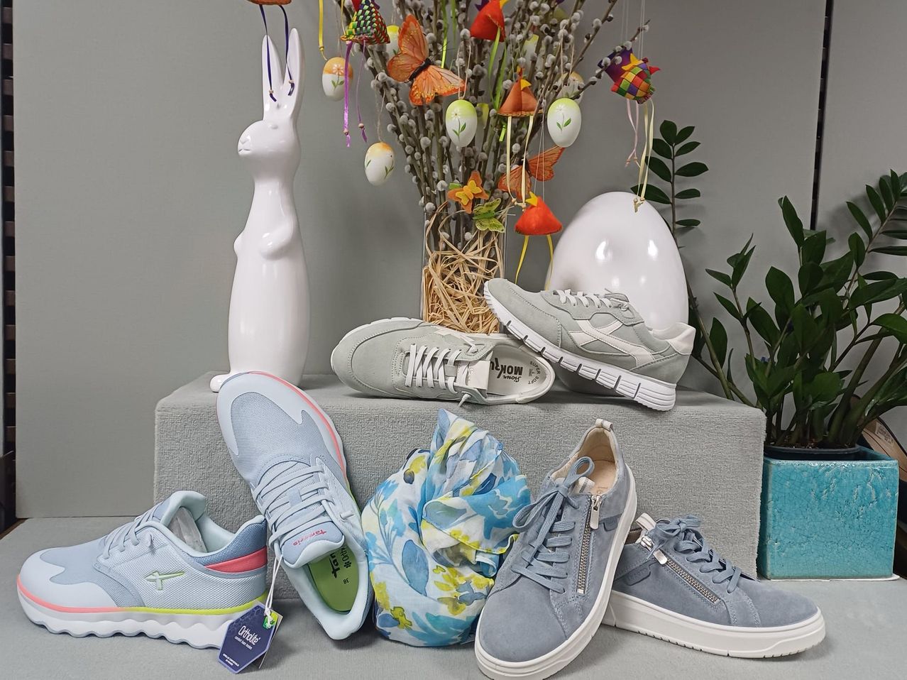 Mehrere Sneaker in verschiedenen Farben und Designs sind auf einer grauen Oberfläche ausgestellt, begleitet von Osterdekorationen wie einem Hasen, Eiern und Schmetterlingen.