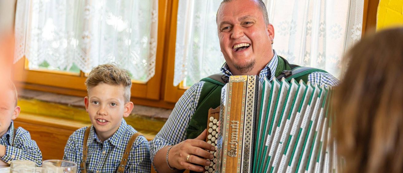 Ein Mann in traditioneller deutscher Kleidung spielt Akkordeon, während er mit einem jungen Jungen am Tisch sitzt.