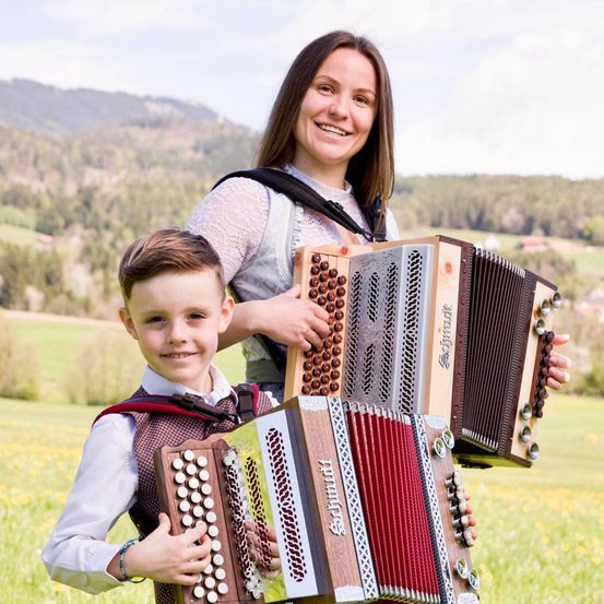 Eine Frau und ein Junge stehen in einem Feld und halten Akkordeons. Die Frau lächelt und der Junge schaut in die Kamera. Hinter ihnen ist eine Bergkette.