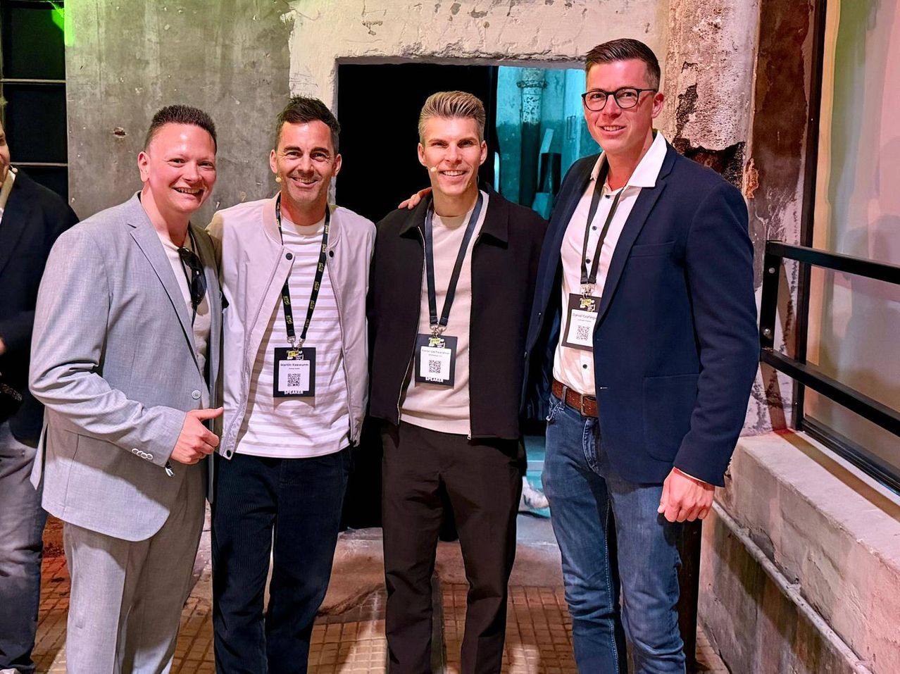 Four men stand together in a well-lit room. They wear IDs with QR codes. Two wear formal attire, and two wear casual clothes. They smile for the camera.