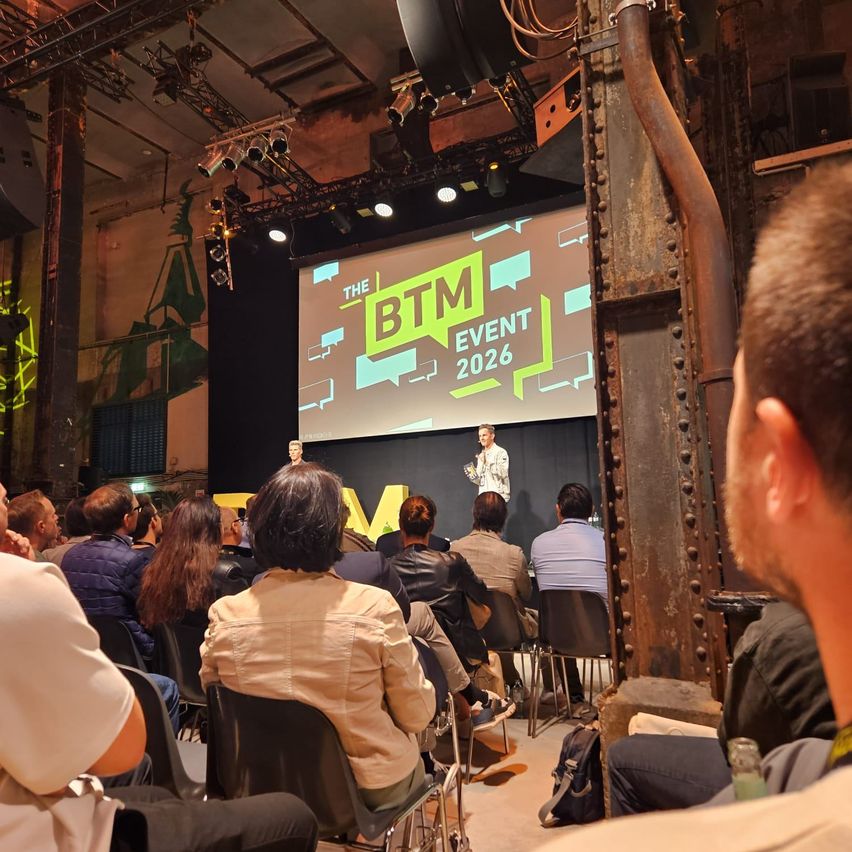 A crowd of people sit in chairs watching a presentation on stage. The screen behind them displays 'The BTM Event 2026'. Two individuals stand on stage, one addressing the audience.