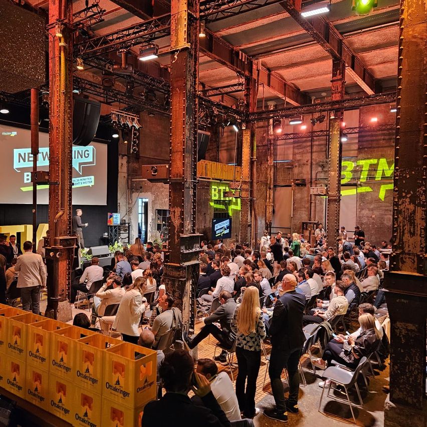 A large indoor event space with an audience seated in rows, a stage with speakers, a screen displaying 'NETWORKING', and yellow 'BTM' signs. The room has a rustic industrial feel with exposed beams.