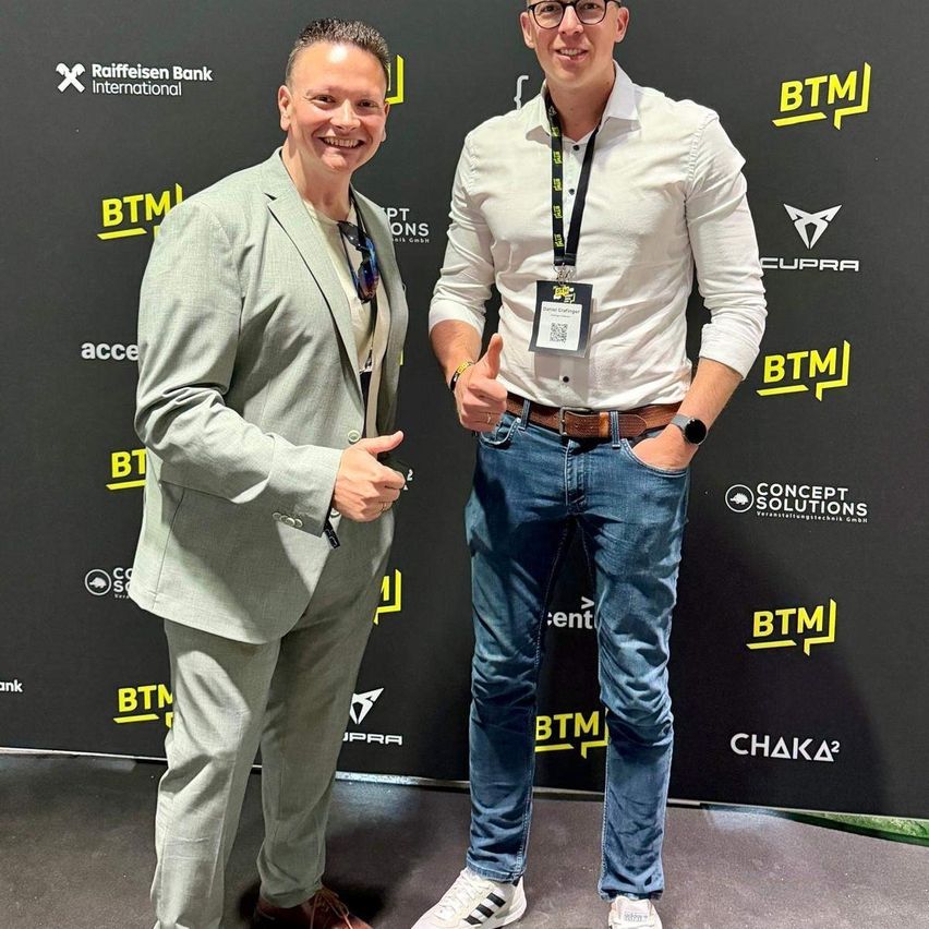 Two men stand close, smiling, in front of a branded wall. One wears a gray suit, the other a white shirt with a name tag. The wall is adorned with logos and text.