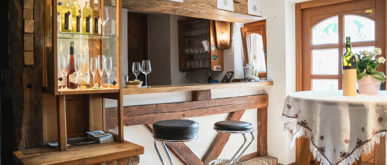 Eine Bar mit zwei schwarzen Barhockern, einem Tisch und einem Regal mit Weingläsern und Flaschen.