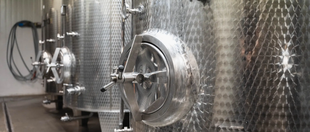 Edelstahl-Weintanks in einem Weinkeller mit mehreren Ventilen und einem offenen Verschluss.