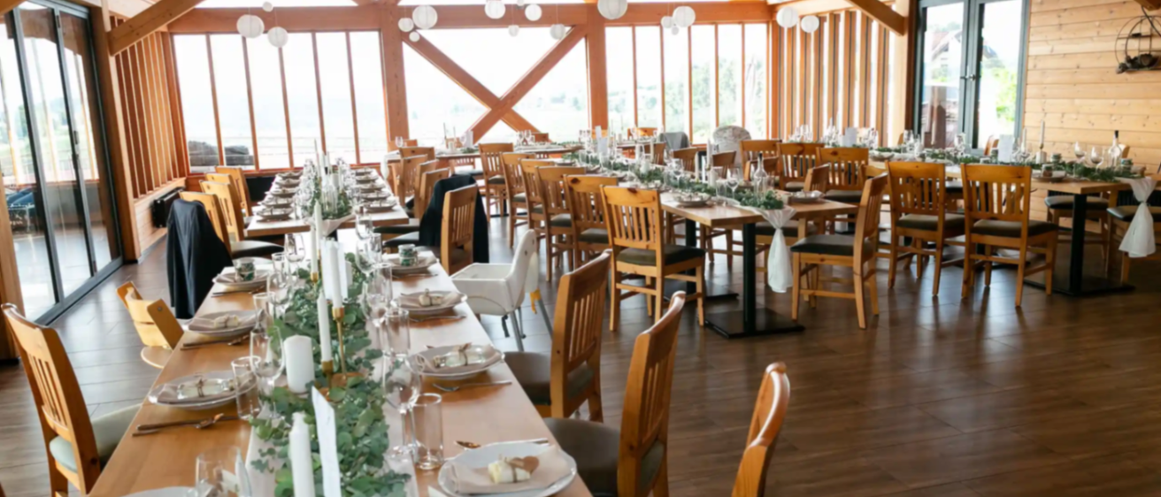 Ein Speisesaal in einem Restaurant mit mehreren Tischen und Stühlern. Ein langer Tisch mit weißen Kerzen und Tellern ist für eine Mahlzeit vorbereitet.