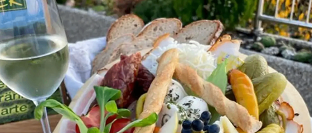 Ein Holztablett gefüllt mit einer Auswahl an Fleisch, Gemüse und Früchten. Das Tablett enthält geschnittenen Schinken, gekochte Eier, Gurkenscheiben und verschiedene Beeren. Ein Glas Weißwein steht nebenan.