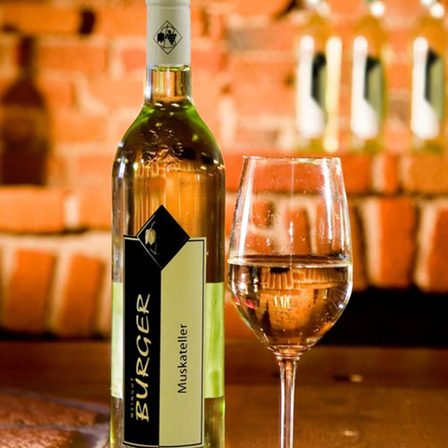 Eine Flasche Burger Muskatteller Wein und ein gefülltes Glas Wein auf einem Tisch mit einer Ziegelwand im Hintergrund.