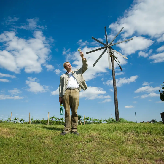 Ein Mann steht auf einem Feld, lächelt und hebt ein Weinglas in die Luft. Im Hintergrund steht eine Windmühle gegen einen blauen Himmel mit verstreuten Wolken.