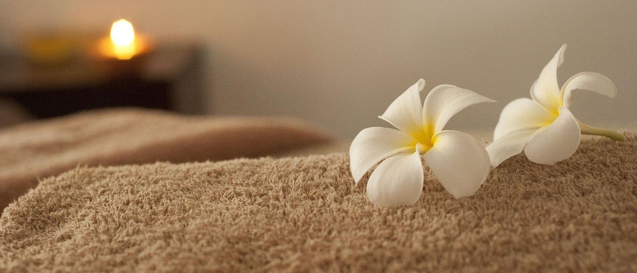 Eine gemütliche Spa-Einrichtung mit weißen Blumen auf einem Handtuch, einer brennenden Kerze im Hintergrund und einem verschwommenen Objekt in der Ferne.