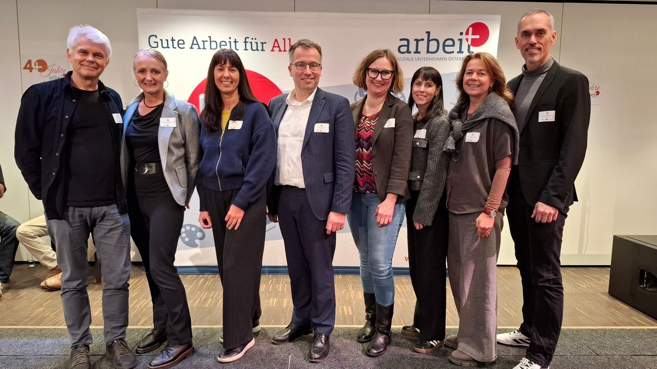 Eine Gruppe von Menschen in Geschäftskleidung steht vor einem Banner mit der Aufschrift 'Gute Arbeit für Alle'. Es sind sechs Menschen im Bild, vier Frauen und zwei Männer.
