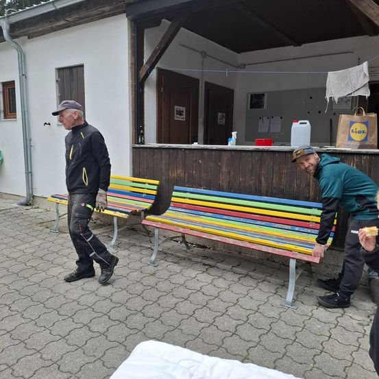 Zwei Männer stehen neben bunten Bänken vor einem Gebäude mit einem Tresen, einem hängenden Handtuch und einer Lidl-Tasche. Ein Mann isst, während der andere lächelt.