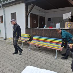 Zwei Männer stehen neben bunten Bänken vor einem Gebäude mit einem Tresen, einem hängenden Handtuch und einer Lidl-Tasche. Ein Mann isst, während der andere lächelt.