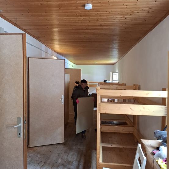 Zwei Personen befinden sich in einem Raum mit holzfarbenen Etagenbetten. Eine Person hält eine Pappe hoch und eine andere Person steht in der Nähe einer Tür. Der Raum hat eine hölzerne Decke und einen Boden.