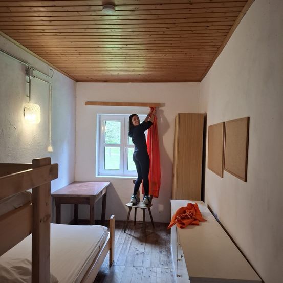 Eine Frau in einem schwarzen Outfit steht auf einem Hocker und hält einen Vorhang in der Nähe eines Fensters in einem Zimmer mit Holzböden. Ein Holzbett, ein Schreibtisch und ein Schrank befinden sich ebenfalls im Raum.