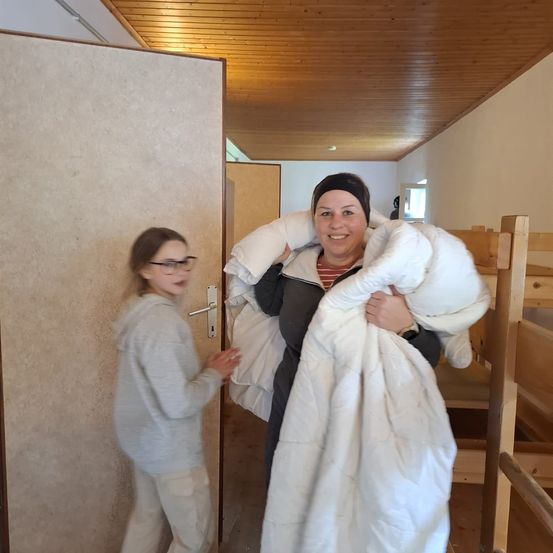 Eine Frau mit einer weißen Decke steht neben einer Tür, während ein Mädchen mit Brille sie öffnet. Sie befinden sich in einem Raum mit einer Holzdecke und einem holzfarbenen Etagenbett.