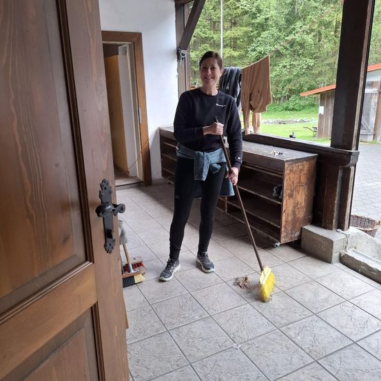 Eine Frau lächelt, während sie den Boden mit einem Besen draußen reinigt. Sie ist in Schwarz gekleidet und hält einen Besen.