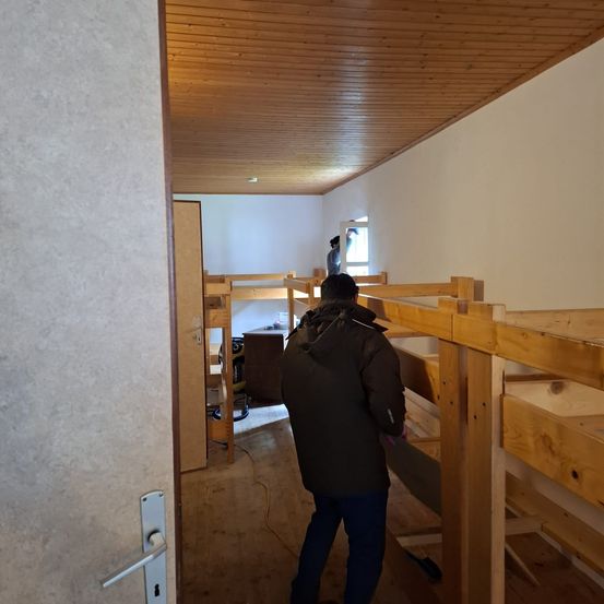 Eine Person steht in einem Raum mit Holzbetten, einem Schreibtisch und einer Person in der Nähe eines Fensters. Der Raum hat eine Holzdecke und weiße Wände.