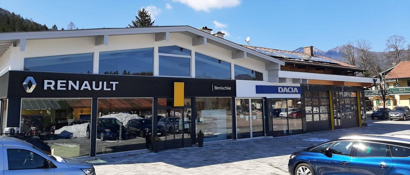 Ein Autohaus mit modernem Design verfügt über eine große Glasfassade, die mehrere Autos zeigt. Im Hintergrund sind ein klarer blauer Himmel und schneebedeckte Berge zu sehen.