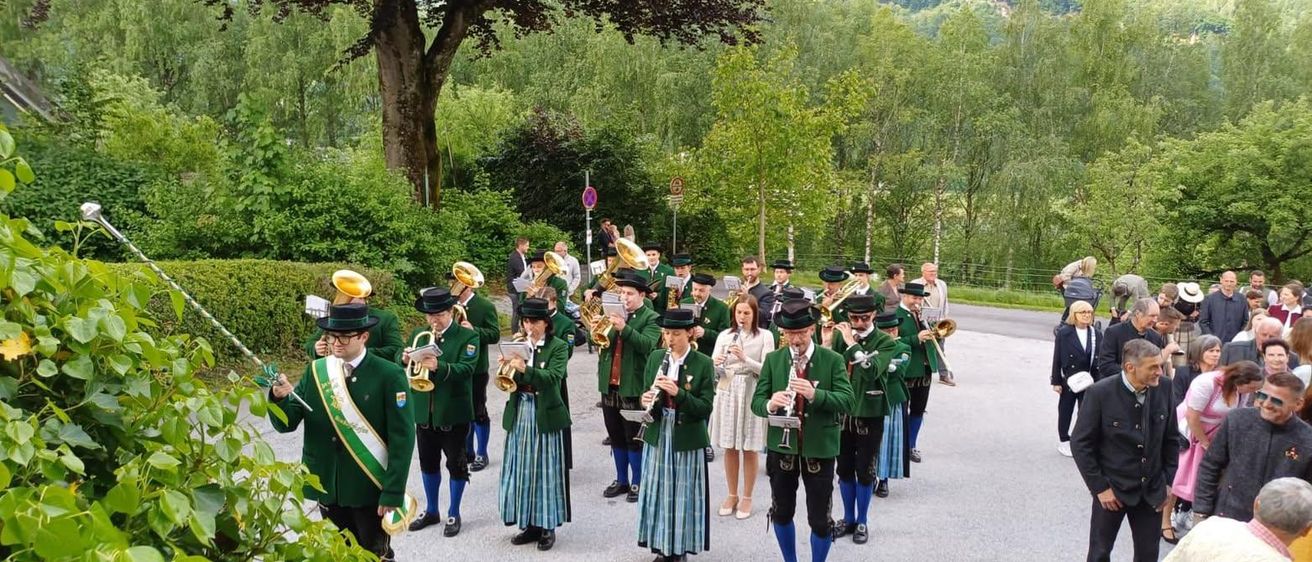 Ein Blasorchester spielt im Freien, die Mitglieder tragen grüne Uniformen und halten verschiedene Musikinstrumente. Eine Frau in einem weißen Kleid steht in der Nähe und beobachtet das Orchester. Die Szene spielt sich in einer üppigen, grünen Umgebung mit Bäumen und Büschen ab.