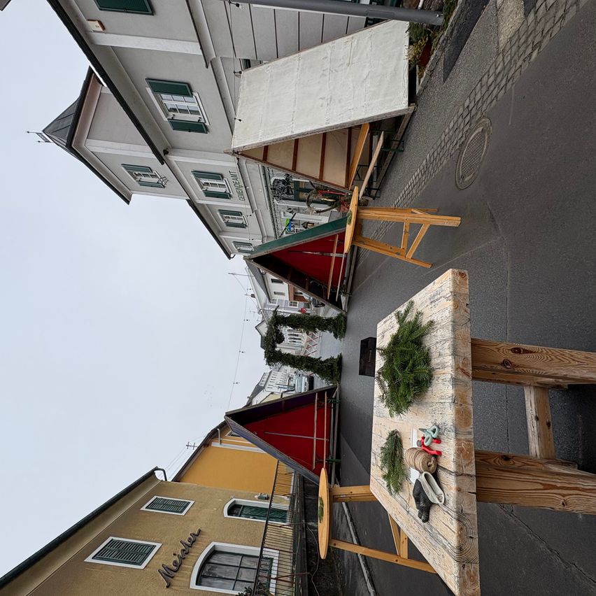 Eine Draufsicht auf eine Straße mit Holztischen, Bänken und einem kleinen Weihnachtsbaum auf einem Tisch. Im Hintergrund sind Gebäude mit Fenstern und einem Schild mit der Aufschrift 'Meier' zu sehen.