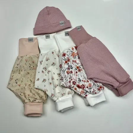 Fünf Babykleidungsstücke, einschließlich einer rosa Mütze und rosa Hosen, sind auf einer weißen Oberfläche ausgebreitet. Die Hosen sind floral und gestrickt.