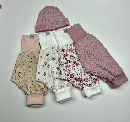 Fünf Babykleidungsstücke, einschließlich einer rosa Mütze und rosa Hosen, sind auf einer weißen Oberfläche ausgebreitet. Die Hosen sind floral und gestrickt.