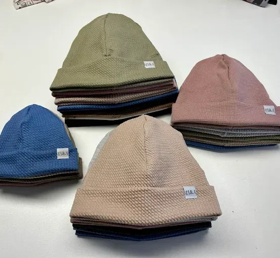 Eine Sammlung von gestrickten Mützen in verschiedenen Farben ist ordentlich auf einer weißen Oberfläche gestapelt, einschließlich Blau-, Rosa- und Beigetönen.