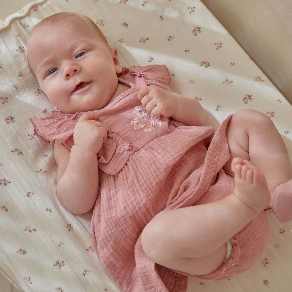 Ein lächelndes Baby liegt auf einer Blumendecke und trägt ein rosa Kleid mit Rüschen und Blumenmuster. Das Baby schaut mit leuchtenden Augen nach oben und hält die Hände hoch.