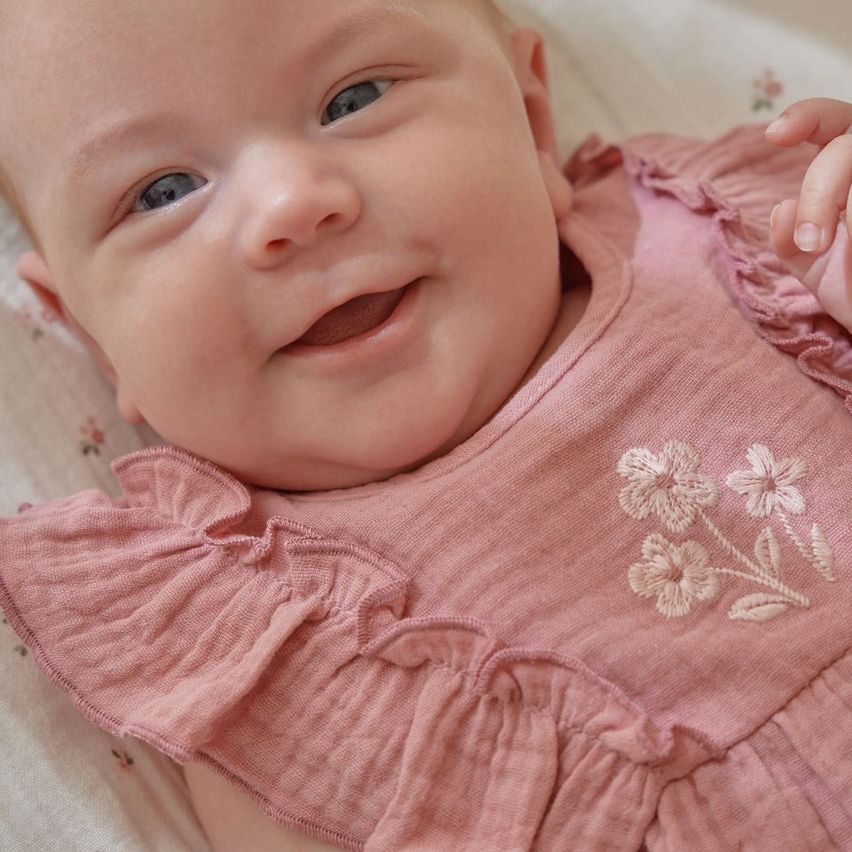 Ein lächelndes Baby liegt auf einer blumengemusterten Decke und trägt ein rosa Kleid mit weißem Blumendesign. Das Baby hat blaue Augen und schaut direkt in die Kamera.