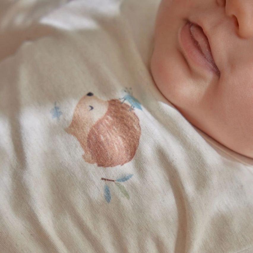 Ein schlafendes Baby liegt auf einem Bett und trägt ein weißes T-Shirt mit einem Igel- und blauen Sternendesign. Das Gesicht des Babys ist leicht verschwommen.