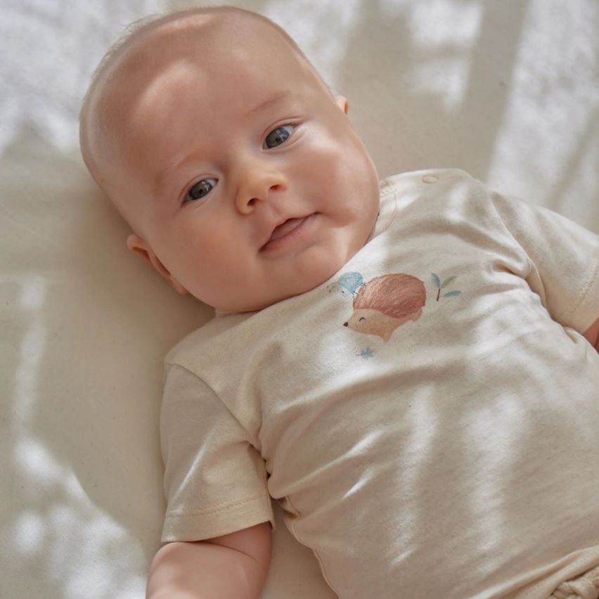 Ein Baby liegt auf einer weißen Decke mit einem weißen T-Shirt, das einen Igel zeigt. Das Baby schaut mit großen Augen umher.