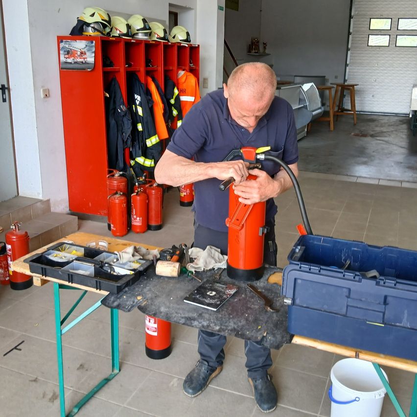 Ein Mann arbeitet an einem Feuerlöscher auf einem Arbeitstisch, mit einem Feuerlöscher-Lagerbereich und Feuerwehrmannsuniformen im Hintergrund.